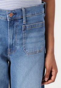 Los jeans de mezclilla azul claro presentan un diseño recto, cierre con botón de cobre y costuras de contraste. El bolsillo trasero es visible a la izquierda.