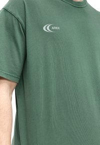 Homme portant un t-shirt vert à manches courtes avec un petit logo blanc "ARKK" et deux lignes courbes sur le haut du côté gauche de la poitrine.
