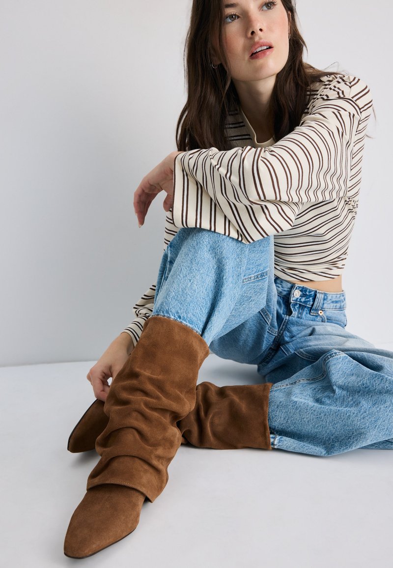Femme aux longs cheveux foncés portant un haut rayé, un jean bleu et des bottes en daim marron décontractées, assise par terre regardant vers le haut.