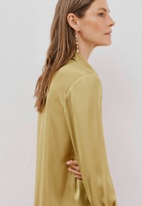 Blusa in satin a maniche lunghe di colore verde oliva, dalla vestibilità rilassata e dalla texture liscia. Presenta un colletto classico e un design minimalista.