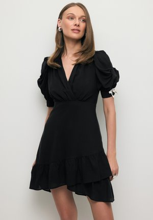 Kobieta o prostych włosach, ubrana w czarną krótką sukienkę z bufiastymi rękawami i marszczonym dołem, stojąca na jasnym, jednolitym tle.