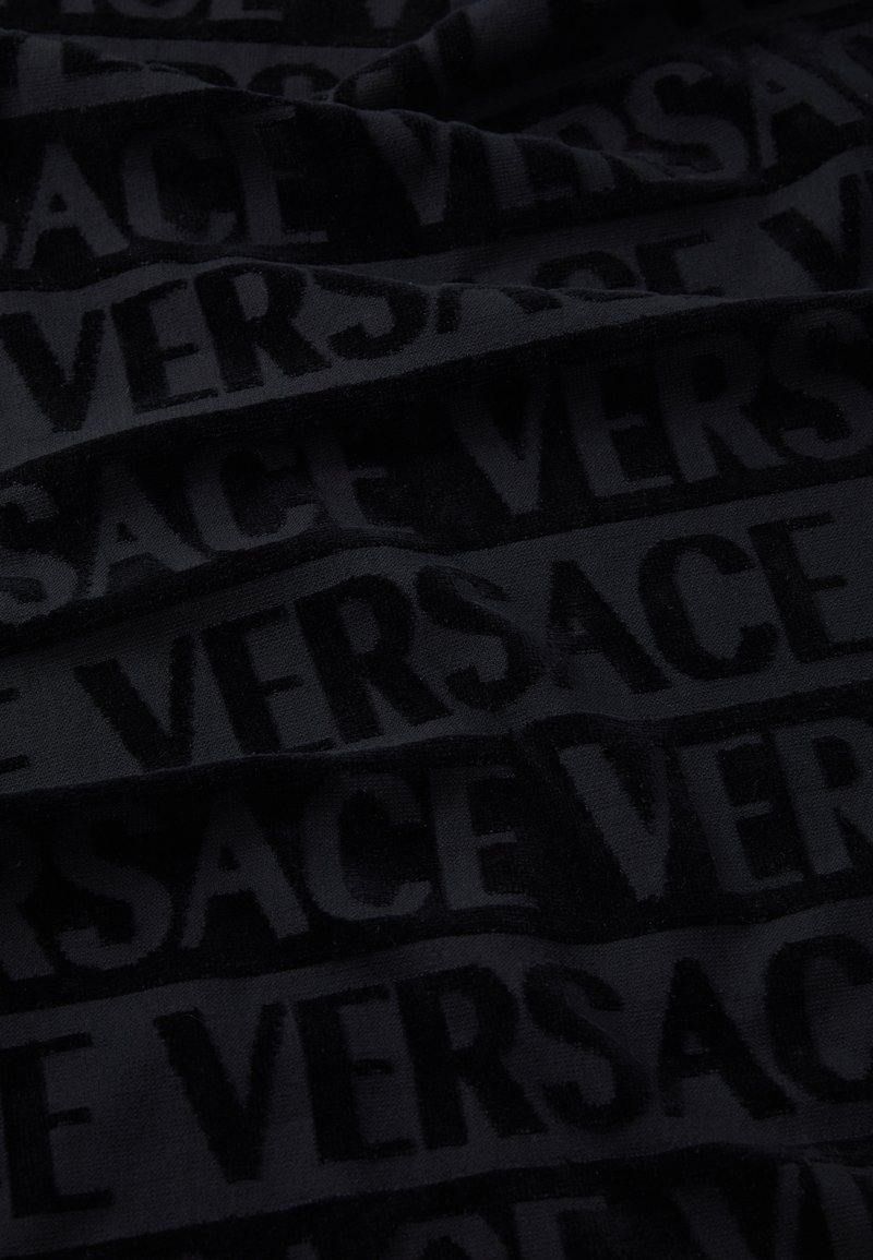 Schwarzer Stoff mit einem wiederholten, strukturierten Muster des Wortes "VERSACE" in verschiedenen Schattierungen, das ein dezentes, aber auffälliges Design erzeugt.