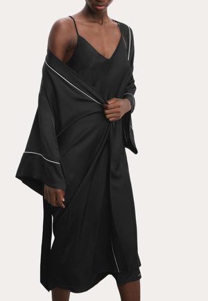Kvinde iført en sort satin natkjole med tynde stropper og en matchende sort kimono med hvide kanter, der holder kimonoen lukket i taljen.
