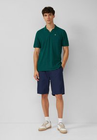 Grünes Baumwollpolo mit einer Tasche, kombiniert mit navy Shorts. Das Modell trägt weiße Sneakers mit beigen Akzenten und weiße Socken. Minimalistisches Design.