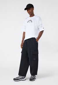 Joven vestido con una camiseta blanca de Champion, pantalones cargo negros, una gorra negra y zapatillas grises, de pie con las manos en los bolsillos sobre un fondo blanco.