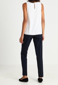 Haut sans manches blanc avec un dos en trou de serrure, texturé avec de petits points. Associé à un pantalon ajusté bleu marine et des chaussures plates noires.