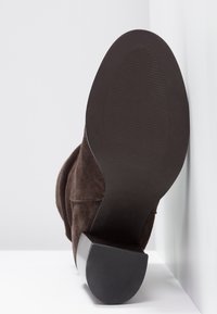 Bota de ante marrón de tacón alto con punta redondeada, superficie texturizada y suela de goma negra sólida. El tacón es grueso y angular.