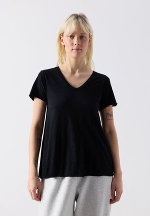 Heledate juustega naine, kellel on seljas avara lõikega must V-kaelusega T-särk ja heledat halli värvi püksid, seisab ühevärvilise hele tausta ees.