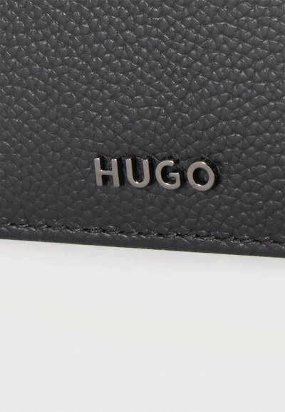 Gros plan d'une surface en cuir noir texturé avec des lettres métalliques argentées formant "HUGO", cousues près du bord inférieur.