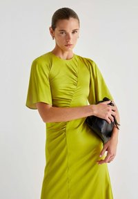 Abito verde brillante con maniche corte e dettagli arricciati, abbinato a una piccola borsa in pelle nera. Tessuto liscio con una silhouette aderente.