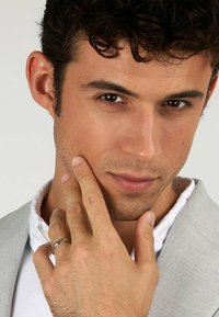 Silverring på handen som rör vid hakan; modell med kort, lockigt mörkt hår bär en ljusgrå blazer över en vit skjorta. Klar hy, fokuserat uttryck.