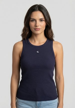 Jeune femme aux longs cheveux noirs portant un débardeur côtelé sans manches bleu marine et un jean bleu clair sur un fond uni.