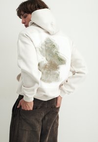 Roomkleurige hoodie met een getextureerd, abstract ontwerp in zachte groentinten en goud aan de achterkant; gecombineerd met donkere corduroybroek.