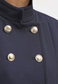 Giacca blu navy con colletto alto, caratterizzata da quattro bottoni argento con loghi in rilievo e un tessuto texturizzato. Design su misura.