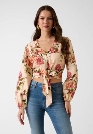 Kobieta ubrana w beżowy, kwiatowy, długi rękaw, krótki top związany w talii, w połączeniu z niebieskimi, wysokim stanem dżinsami na jednolitym tle.