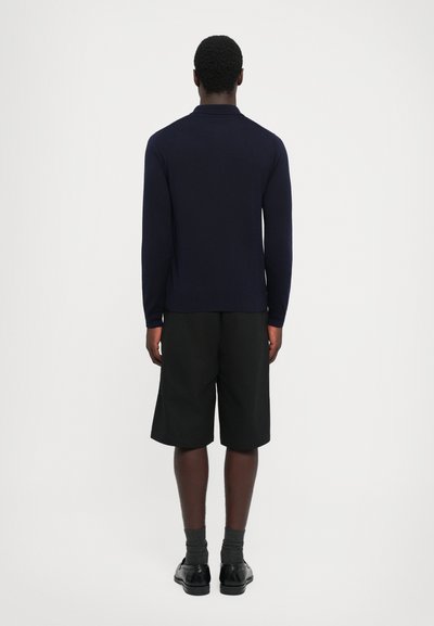 Mörkblå långärmad polo med krage, kombinerad med svarta shorts och mörka skor, sedd bakifrån mot en vit bakgrund.