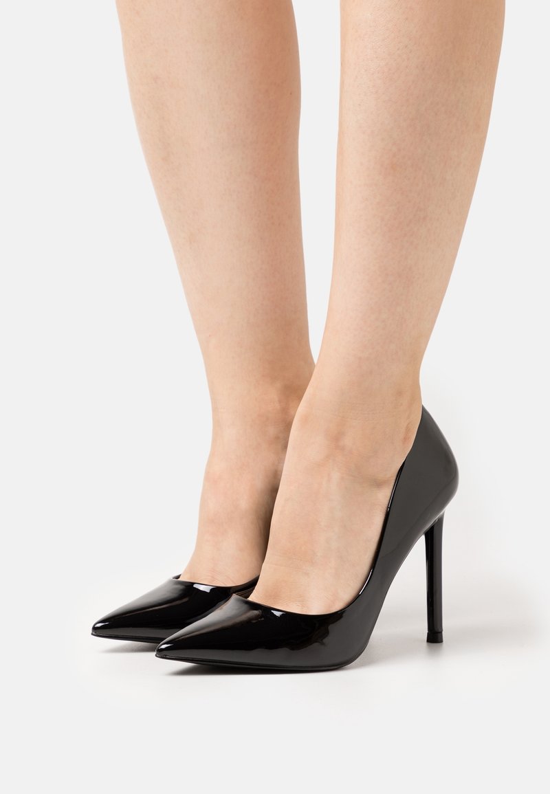Jambes portant des escarpins pointus en cuir verni noir avec des talons aiguilles fins sur fond blanc.