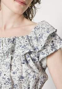 Top com padrão floral em tecido leve, com mangas com volantes, decote quadrado e flores azuis sobre um fundo branco.