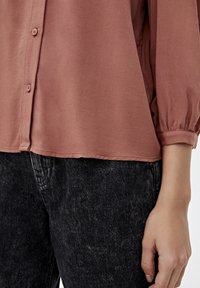 Rose-colored, lightweight blouse with a button-up design and gathered sleeve cuffs, paired with dark, textured denim jeans.