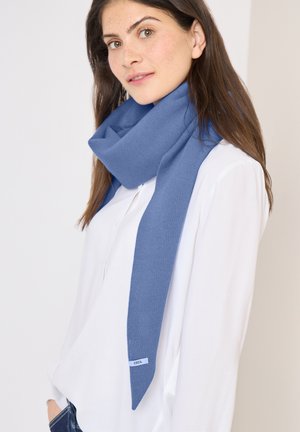 Jeune femme aux longs cheveux bruns portant une écharpe bleue douce sur un chemisier blanc, debout devant un fond neutre.