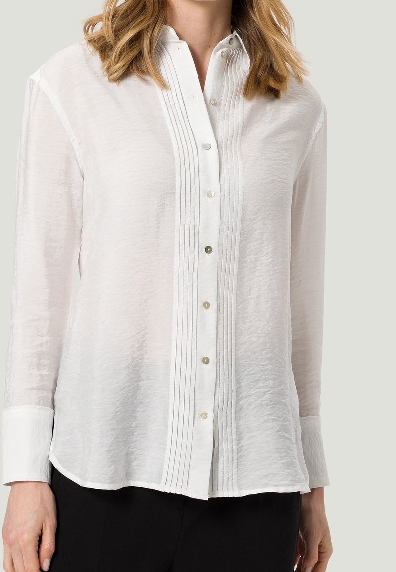 Femme portant un chemisier blanc à manches longues boutonné avec des plis verticaux le long de la ligne de boutons et un pantalon noir.