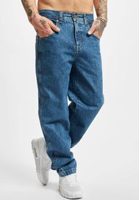 Jeans de mezclilla azul claro con un ajuste relajado, que presentan bolsillos frontales, costuras visibles y un cierre de botón. Llevados con zapatillas blancas.