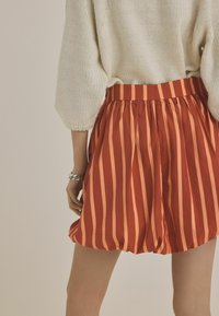 Pantaloni scurți în dungi roșii și portocalii, cu un talie elastică și o croială lejeră, confecționați dintr-un material ușor, asortați cu un pulover alb din tricot.