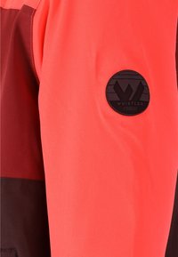 Rote und burgunderfarbene Oberbekleidung mit einer glatten Textur. Ein schwarzes, rundes Logo-Patch mit der Aufschrift "WHISTLER" ist am Ärmel zu sehen.