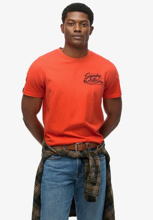 Rød bomulds t-shirt med rund hals, korte ærmer og et sort grafisk print på brystet. Blå jeans og en grønternet skjorte bundet om taljen.