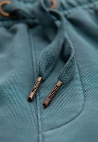 Tirets de capuche teal avec embouts en métal bronze. Le texte sur les embouts lit "DIDIMBRE." Tissu lisse avec des coutures visibles et des détails de texture.
