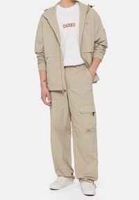 Béžová lehká bunda s kapucí na stahování, doplněná bílým tričkem s nápisem "DICKIES" oranžovým písmem a sladěnými béžovými kalhotami typu cargo.