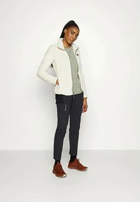 Biała polarowa kurtka na szarym topie, połączona z ciemnymi spodniami. Czekoladowe sneakersy; stoi na jednolitym tle.