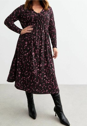 Žena v černých midi šatech s dlouhým rukávem a růžovým abstraktním vzorem, která stojí s rukou v bok a má na nohou černé lodičky s jehlovým podpatkem a špičatou špičkou.