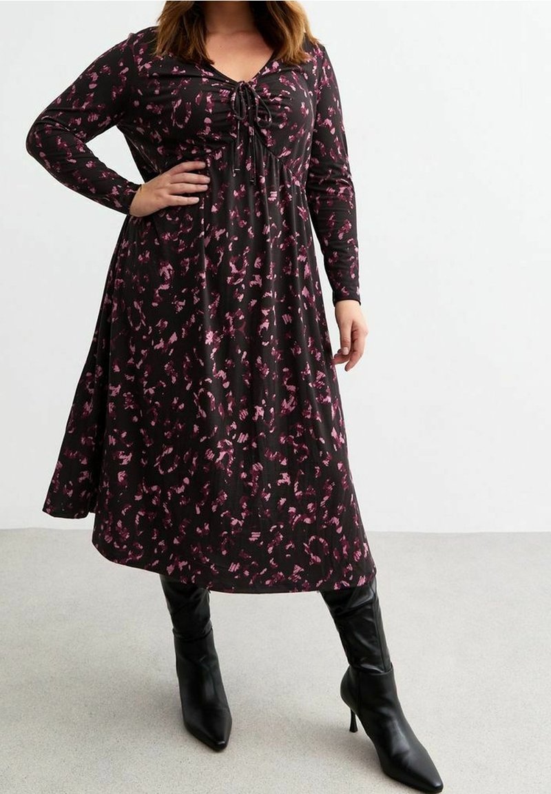 Femme portant une robe midi noire à manches longues avec un imprimé abstrait rose et des bottines noires à talons pointues, debout avec la main sur la hanche.