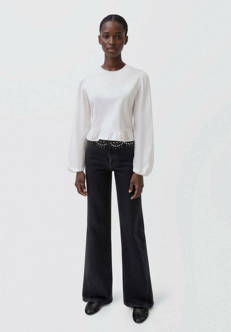 Blusa bianca a maniche lunghe con orlo arricciato abbinata a jeans neri a zampa d'elefante con dettagli borchiati e scarpe nere slip-on.