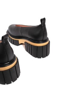 Black leather shoes featuring a rounded toe, chunky black rubber sole, and a wooden accent on the heel. Back loop for easy wear.