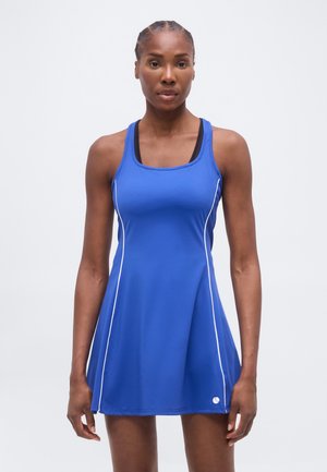 Γυναίκα όρθια, φοράει αμάνικο μπλε αθλητικό φόρεμα με λευκές ραφές, κοιτάζοντας προς τα εμπρός σε απλό φωτεινό φόντο.