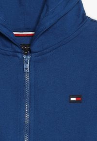 Felpa blu con zip realizzata in tessuto morbido, caratterizzata da un alto colletto, una patch con logo del marchio e strisce rosse e bianche a contrasto all'interno del colletto.
