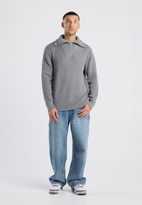 Hombre de pie vistiendo un suéter de manga larga gris con cuello de cremallera, unos jeans holgados de color azul claro y zapatillas blancas, frente a un fondo blanco.