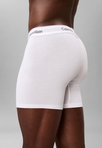 Męski model noszący białe bokserki Calvin Klein na jednolitym szarym tle, pokazujący widok z boku i z tyłu.