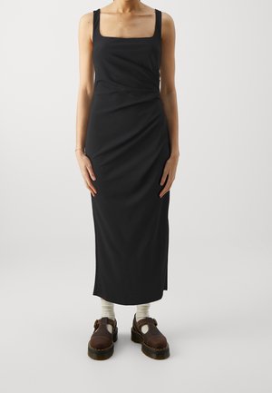 Schwarzes ärmelloses Kleid mit quadratischem Ausschnitt, figurbetonter Silhouette und gerafftem Detail an der Taille. Getragen mit braunen klobigen Schuhen und weißen Socken.