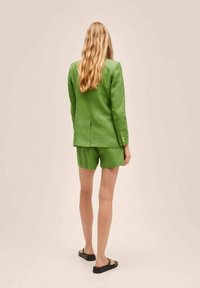 Blazer verde in lino con pantaloni corti abbinati. Il blazer presenta una vestibilità sartoriale e bottoni decorativi. Indossato con sandali neri slip-on.