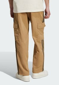 Pantalon cargo beige avec deux poches latérales, bandes noires sur les côtés et texture légère. Porté avec des baskets blanches, affichant une coupe décontractée.