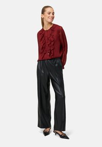 Bordeauxroze blouse met ruches en knopen, gecombineerd met zwarte wijde broek van nep leer en puntige kitten heels, tegen een effen achtergrond.