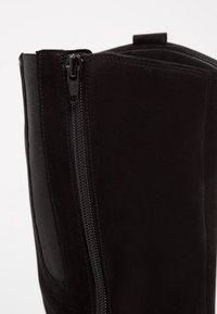 Gros plan sur la tige d'une botte en daim noir, mettant en avant une fermeture éclair verticale YKK et un panneau latéral élastique pour un ajustement flexible.