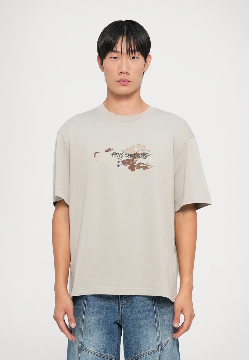 Lys beige oversized t-skjorte laget av bomull, med et fargerikt trykt design på forsiden som har abstrakte mønstre og tekst. Korte ermer.