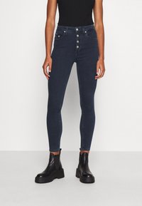 Mörktvättade högmidjade jeans med en figurnära silhuett, femknappsstängning och flera fickor. Matchas med svarta kraftiga ankelstövlar.