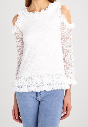 Witte kanten blouse met open schouders, ruches langs de halslijn en zoom, gedragen met lichtblauwe spijkerbroek, armen ontspannen langs het lichaam.