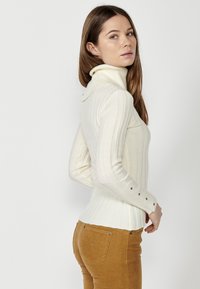 Cremfarbener gerippter Rollkragenpullover mit Knöpfen an den Bündchen und einer schmalen Passform, kombiniert mit senfgelben Cordhosen für einen Kontrast.