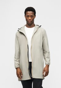 Νέος άντρας που φοράει ανοιχτό γκρι αδιάβροχο με κουκούλα πάνω από λευκό πουκάμισο και μαύρο παντελόνι, στέκεται μπροστά σε απλό ανοιχτό υπόβαθρο.